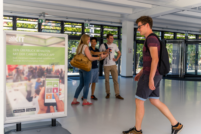 Studierende unterhalten sich im Hintergrund und ein Studierender läuft vorbei an dem Aufsteller für Campuswerbung